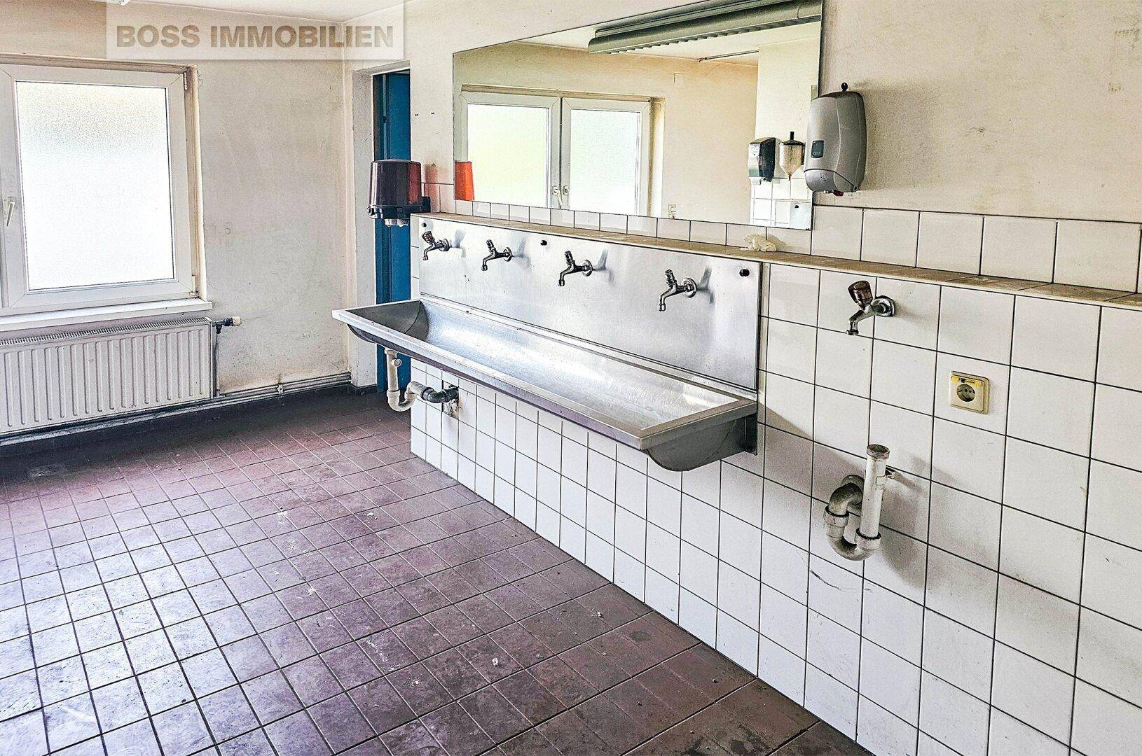 Geräumiger Waschraum mit langem Edelstahlwaschbecken, gefliesten Wänden und Fenster.