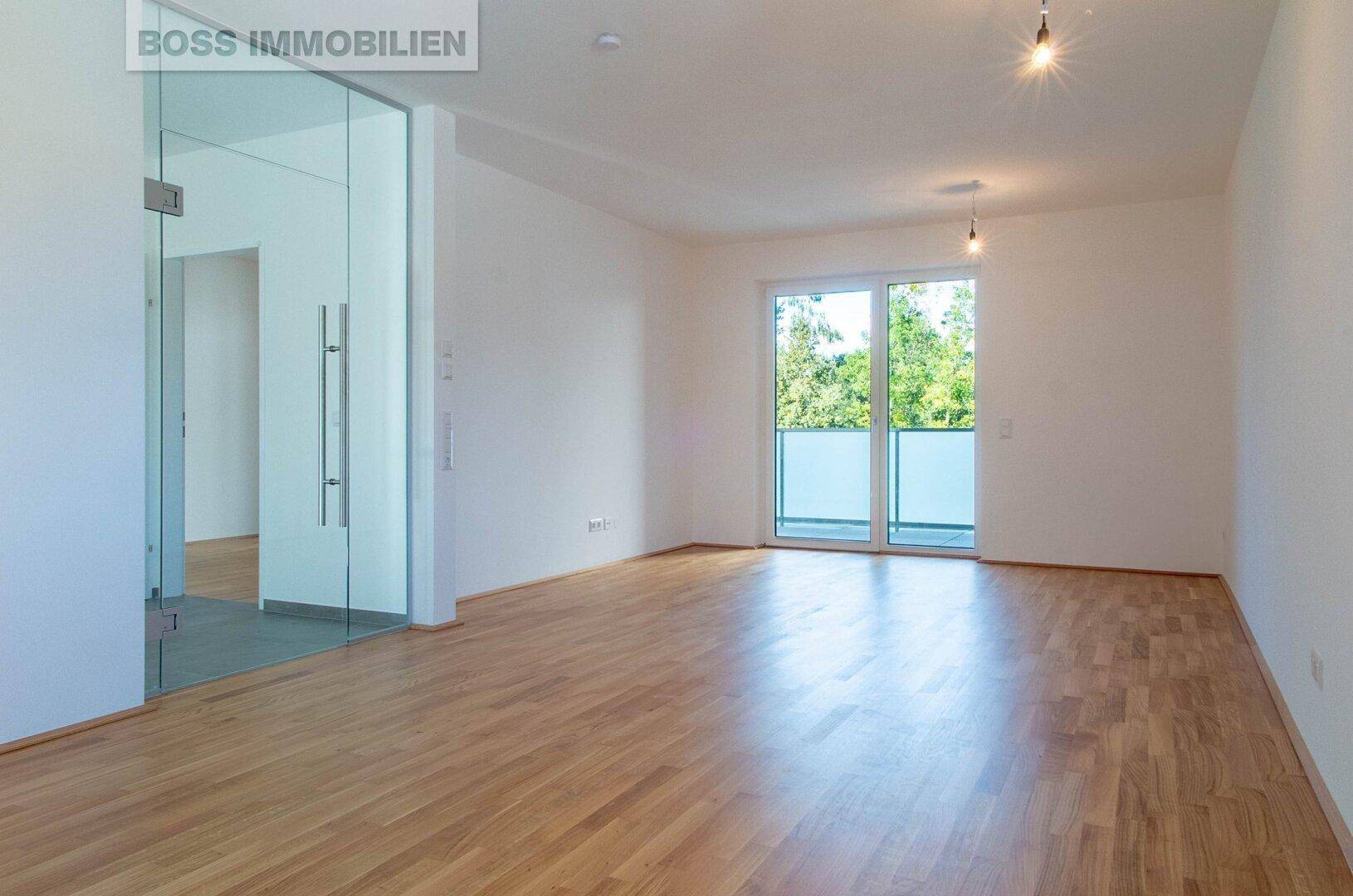 Geräumiger, leerer Raum mit hellem Parkettboden, Glaswand und Zugang zu einem Balkon.