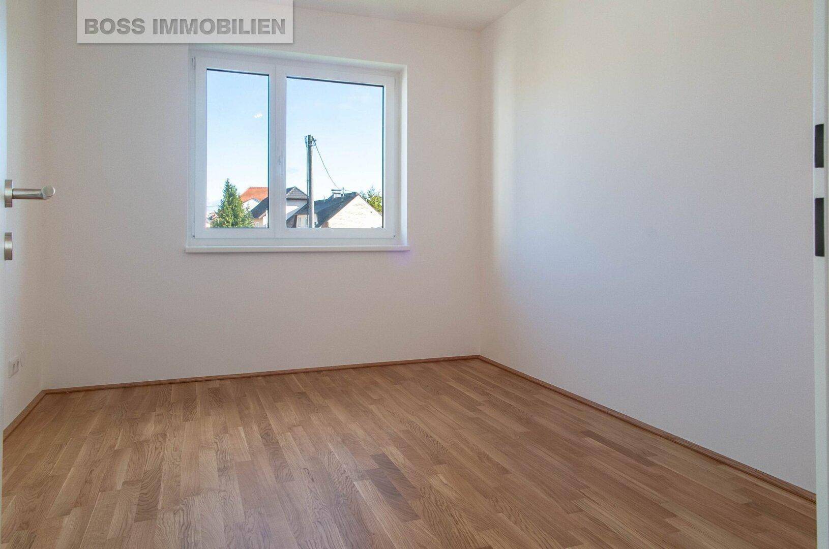 Ruhiges Schlafzimmer mit hellem Parkettboden und einem Fenster mit Blick ins Grüne.