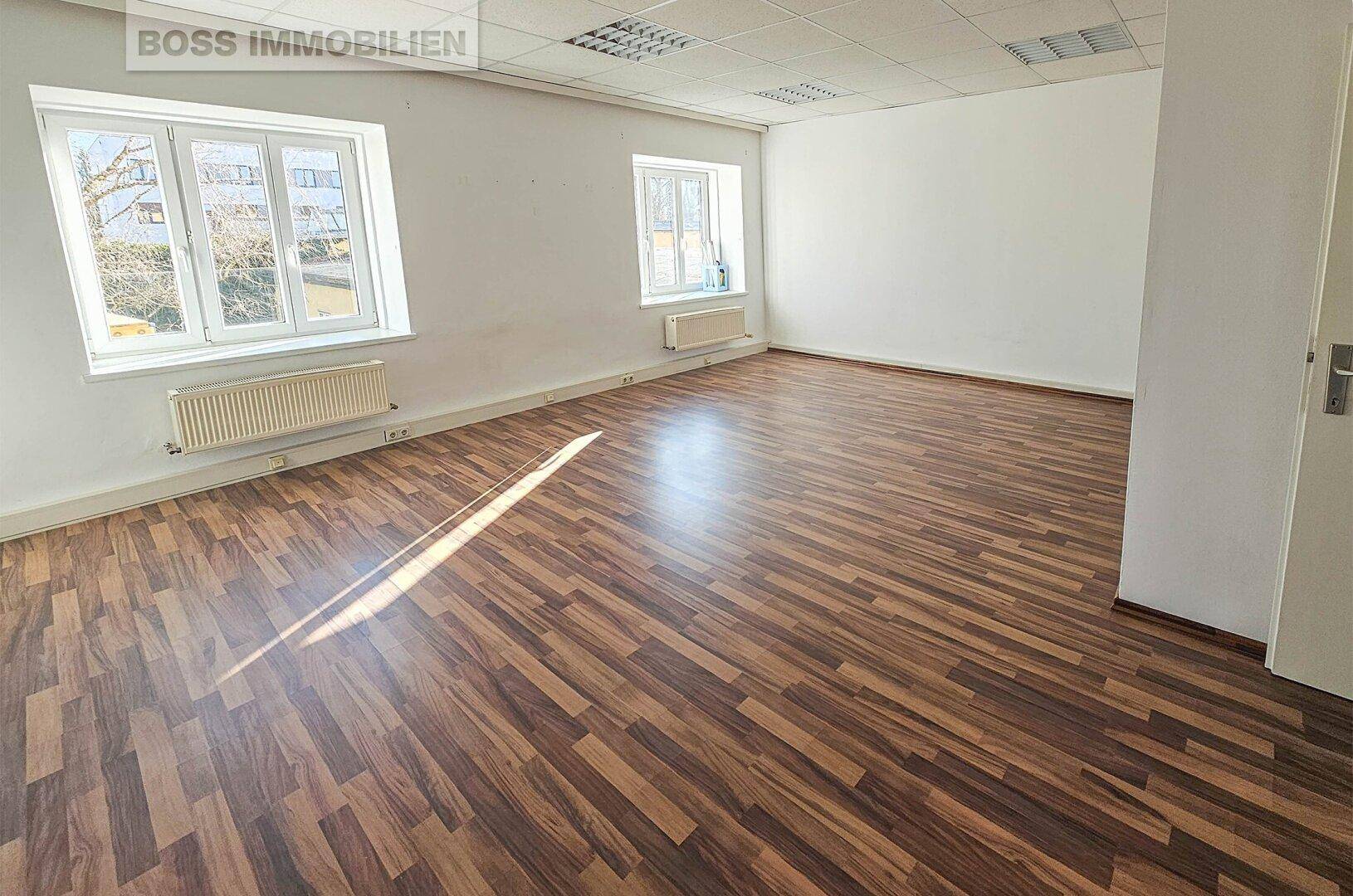 Heller Büroraum mit Laminatboden und großen Fenstern, die viel Tageslicht hereinlassen.