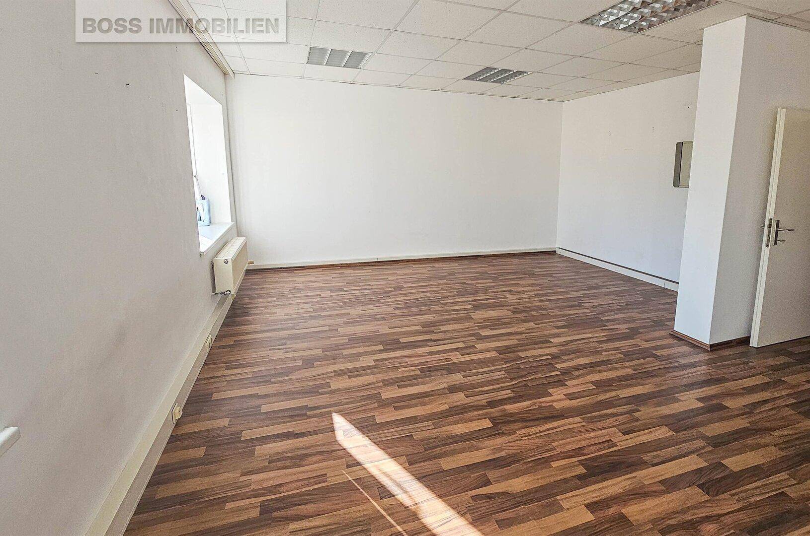 Geräumiger Bürobereich mit dunklem Laminatboden und schlichten weißen Wänden.