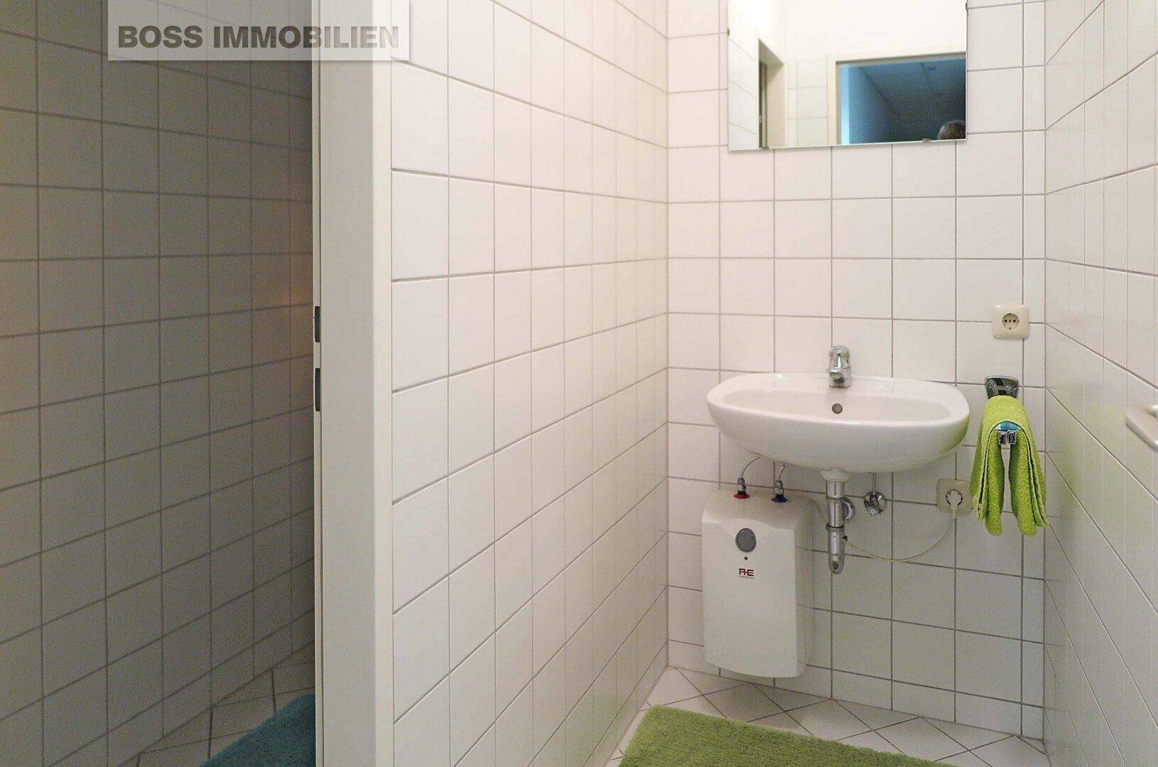 Helles Badezimmer mit weißen Fliesen, Waschbecken und einem kleinen Warmwasserbereiter.