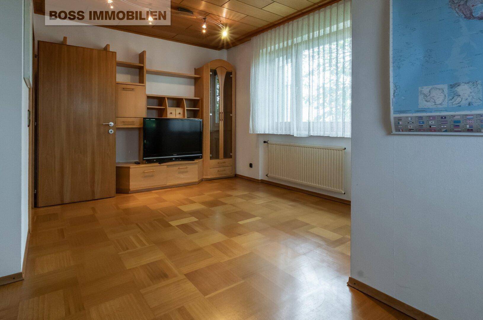 Zimmer mit Parkettboden, Holzmöbeln und Fenster mit Blick ins Grüne.