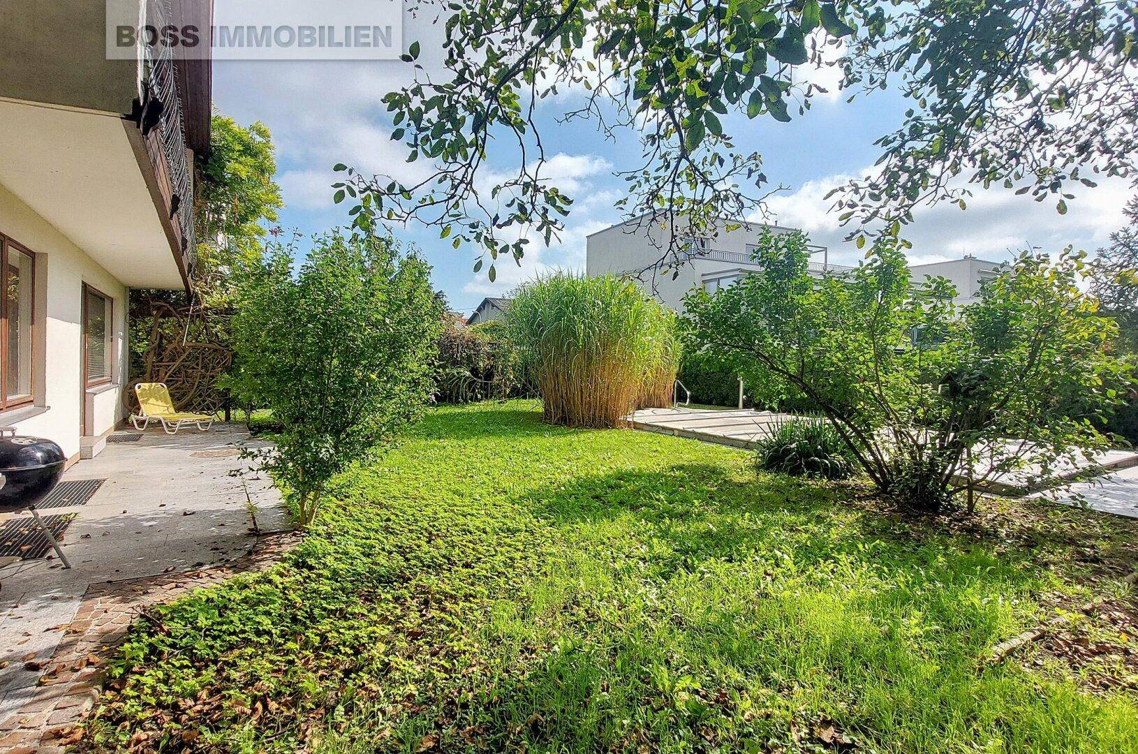 Weitläufiger Garten mit Rasen, Sträuchern und einer Terrasse im Hintergrund.