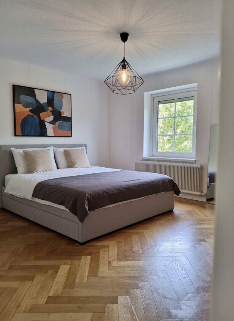 Helles Schlafzimmer mit Parkettboden, großem Bett und modernem Deckenlicht.