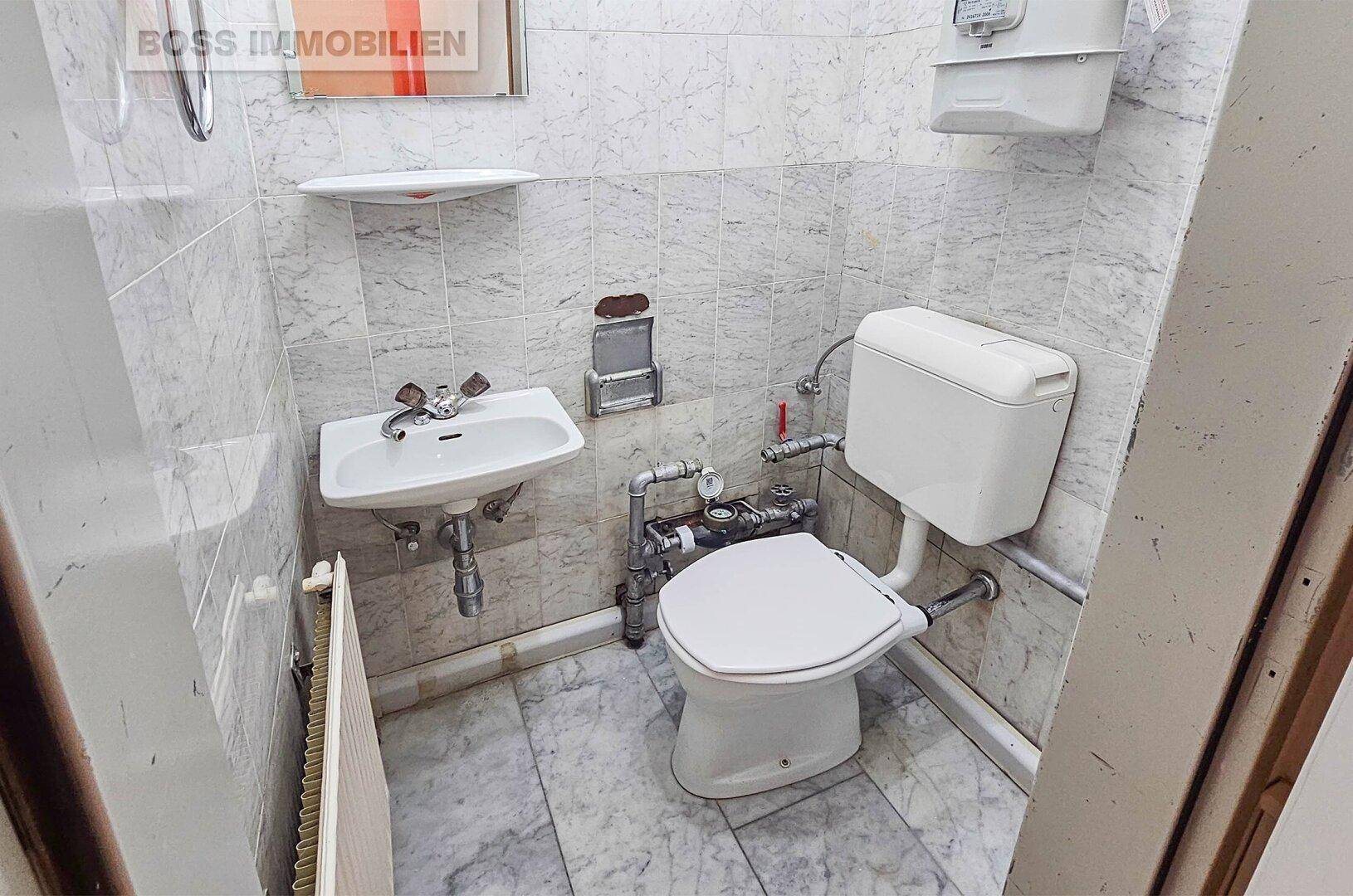 Einfache Toilette mit Waschbecken, WC und gefliesten Wänden und Boden in Marmoroptik.