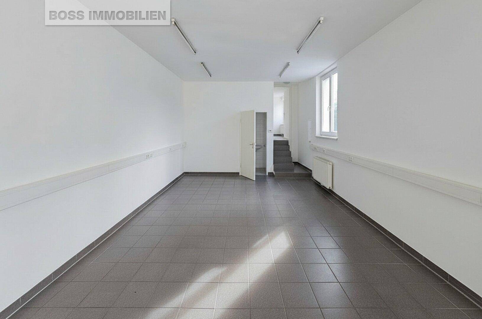 Leerer Gewerberaum mit Fliesenboden, weißen Wänden und Zugang zu einer Treppe, gut beleuchtet.