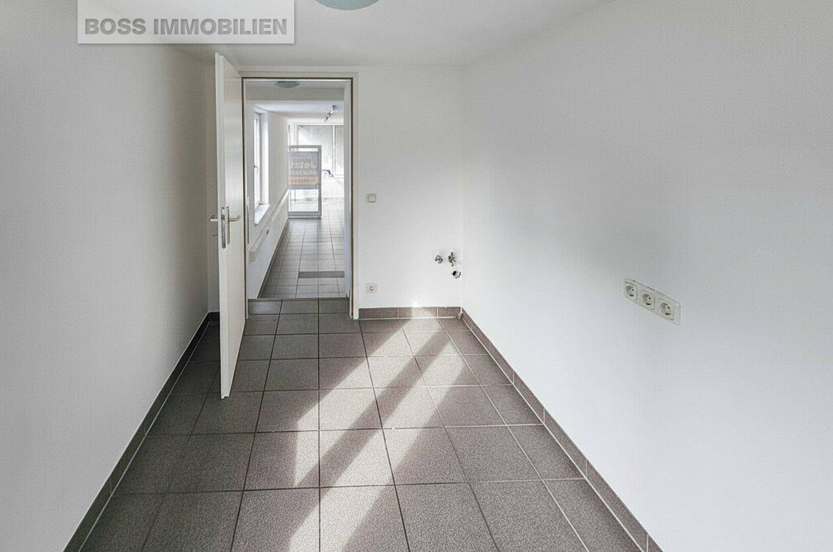 Leerer Raum mit Fliesenboden und weißen Wänden, mit sichtbaren Anschlüssen und Zugang zu einem Flur.