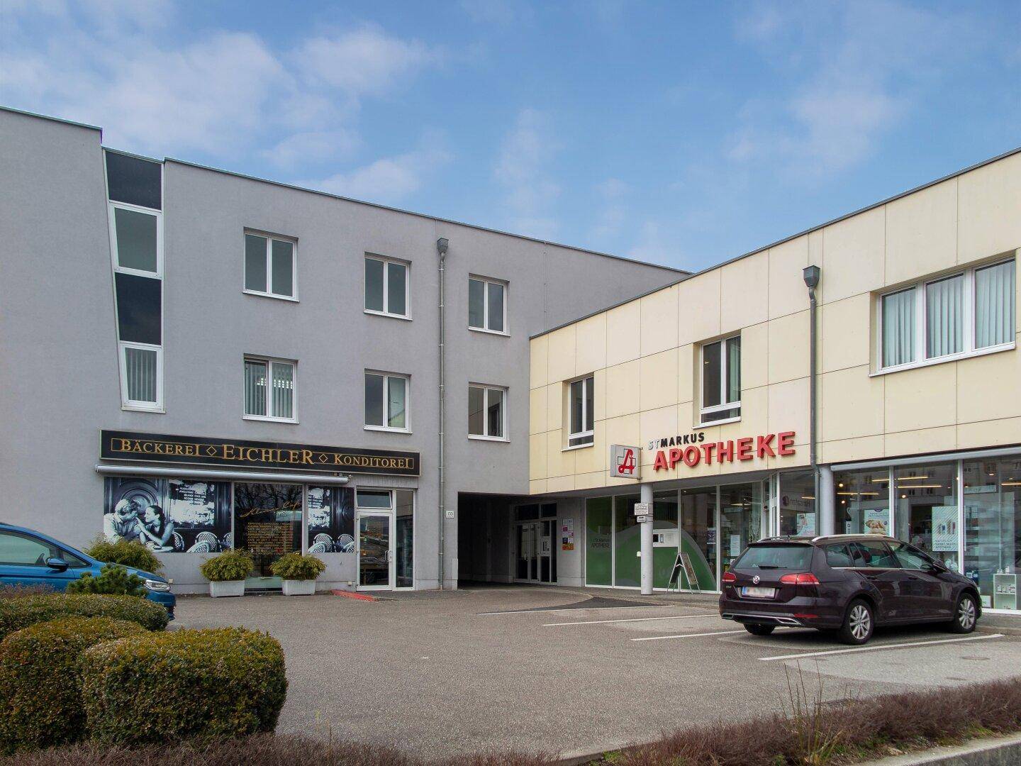Außenansicht eines modernen Geschäftsgebäudes mit Bäckerei und Apotheke sowie Parkplätzen davor.