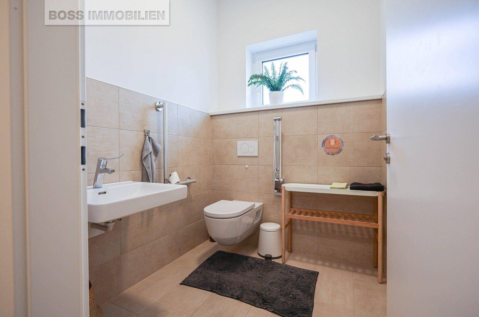 Helles Badezimmer mit beigefarbenen Fliesen, wandmontierter Toilette und Haltegriffen für Barrierefreiheit.