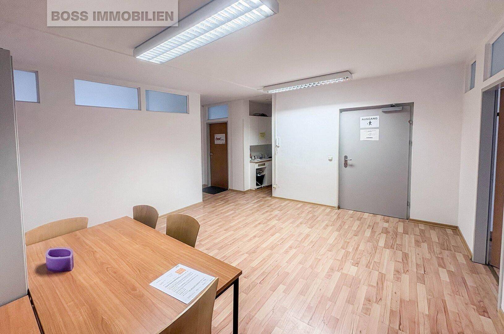 Offener Bürobereich mit Esstisch, Stühlen und einer kleinen Teeküche im Hintergrund, gut beleuchtet.