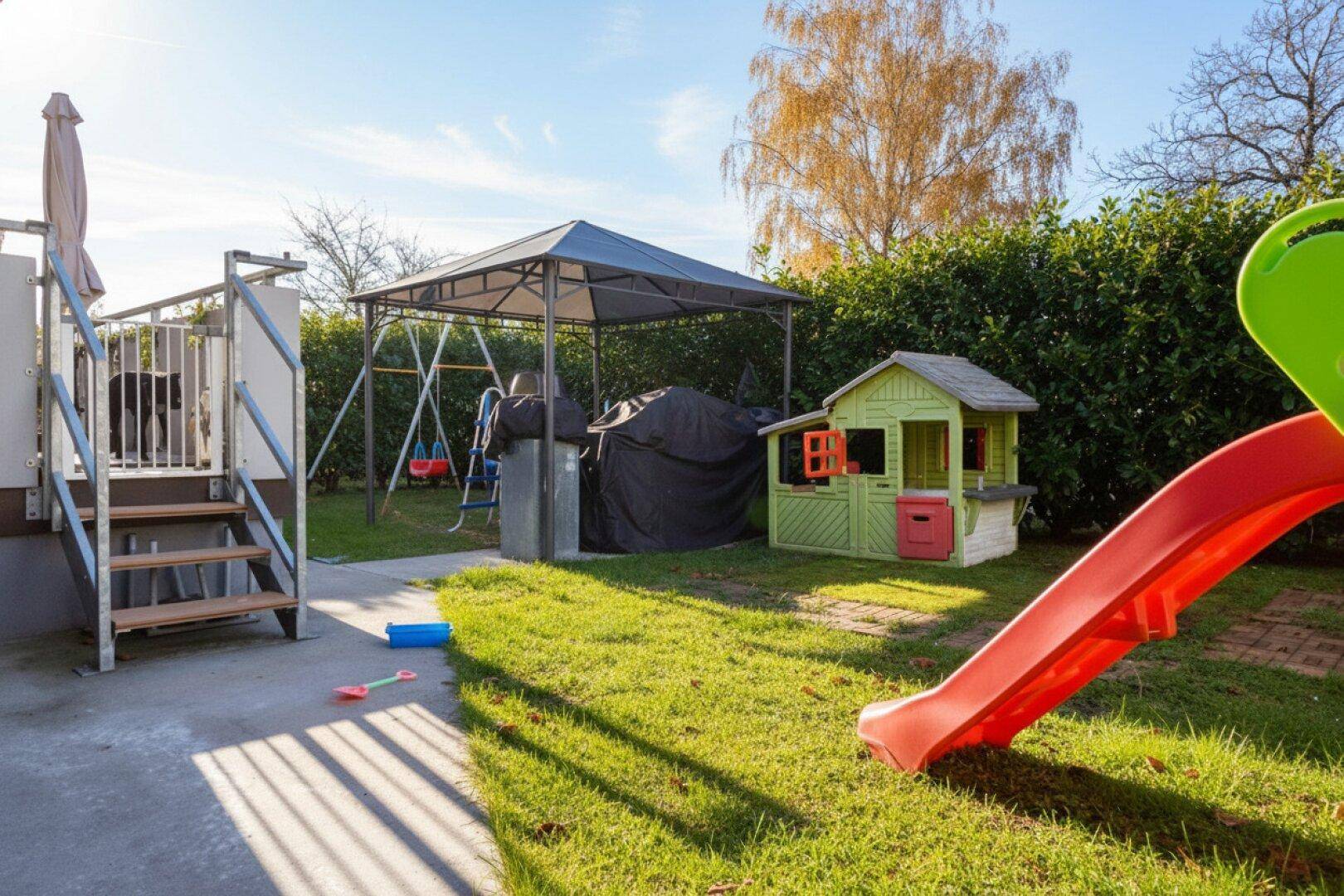 Sonniger Garten mit Spielhaus, Rutsche und Schaukel für Kinder.