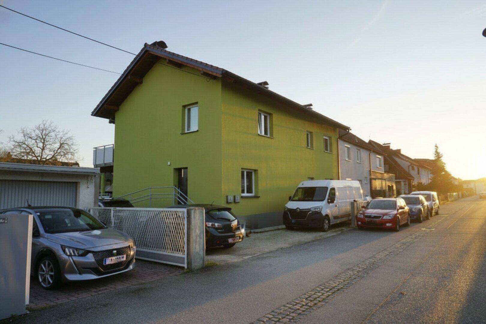 Grüne Hausfassade mit Satteldach und Parkmöglichkeiten an der Straße bei Sonnenuntergang.