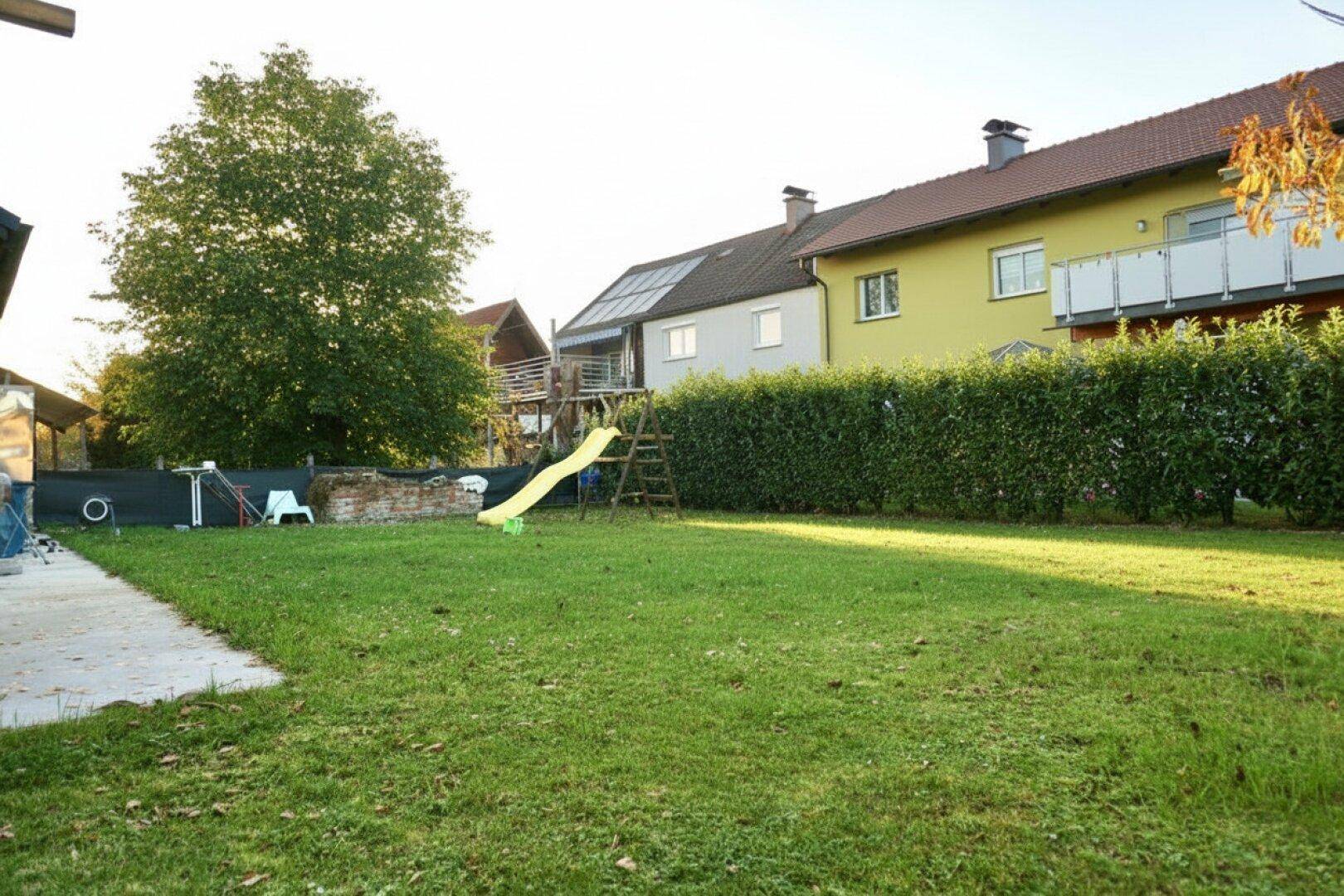Weitläufiger Garten mit grüner Hecke, Rasenfläche und Kinderrutsche im Sonnenlicht.