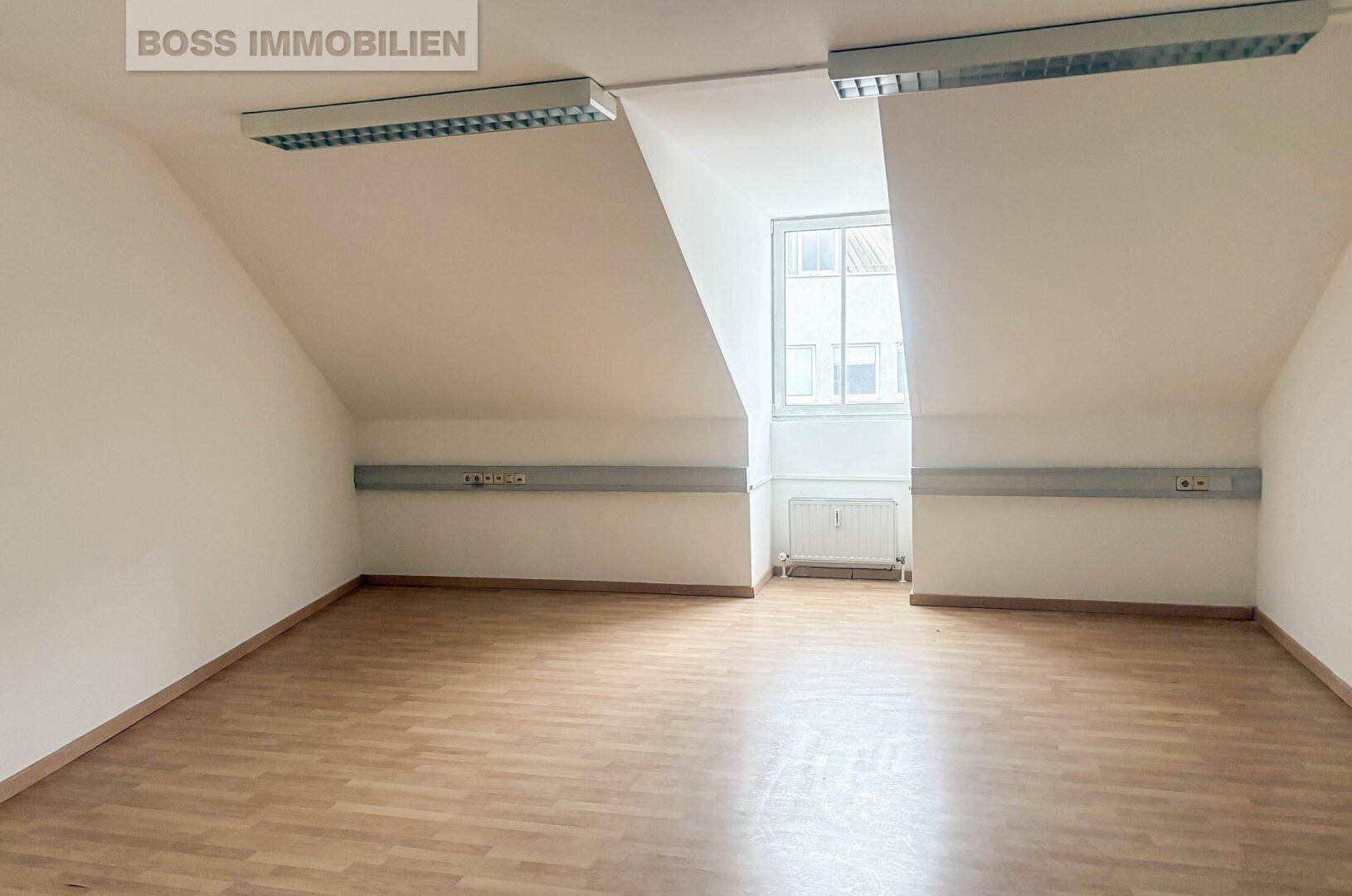 Leerer Büroraum mit Dachschrägen, Laminatboden und einem Fenster, das viel natürliches Licht bietet.