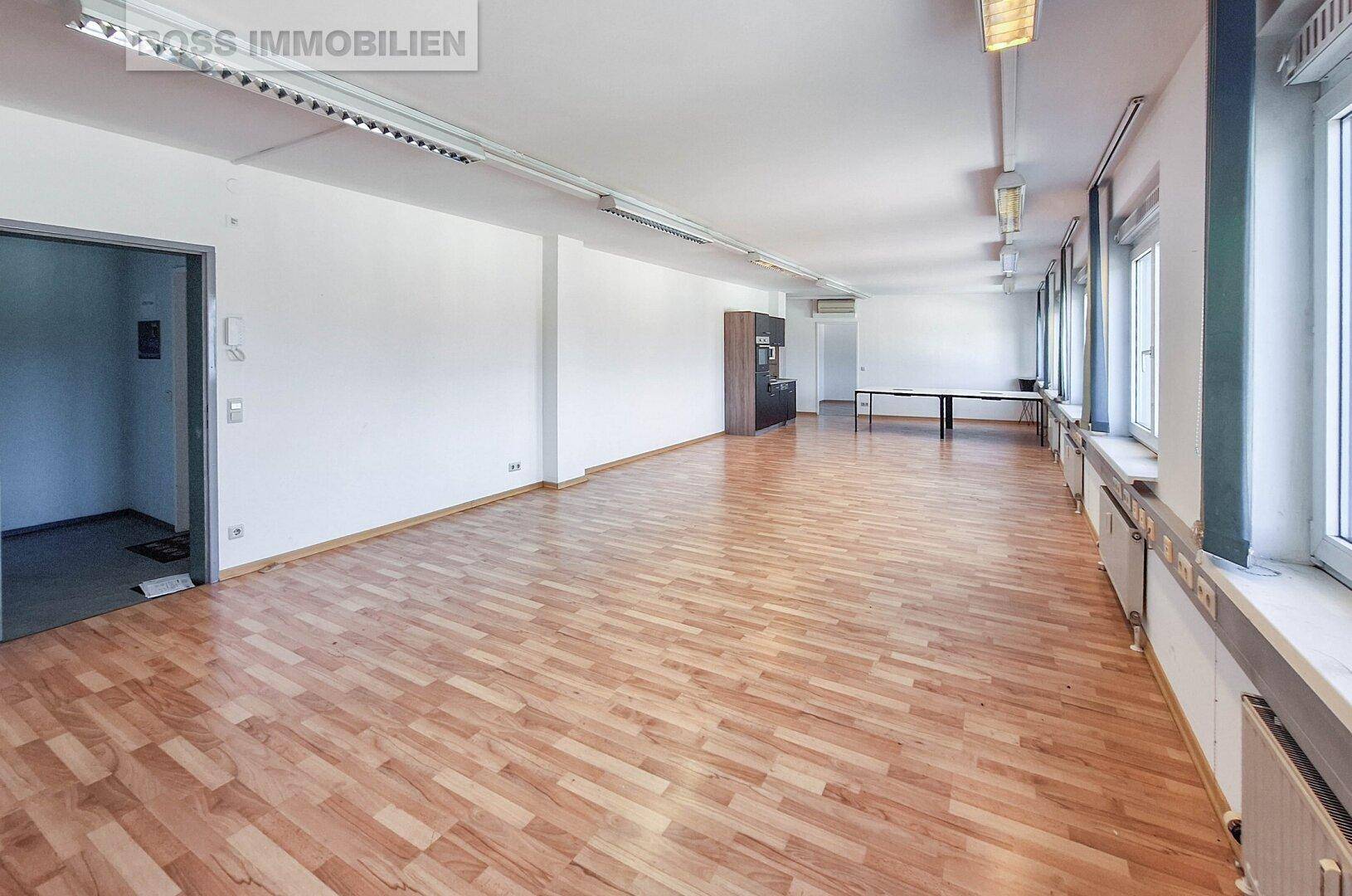 Langer Büroraum mit Laminatboden, weißen Wänden und einem Eingang zu einem Nebenraum.