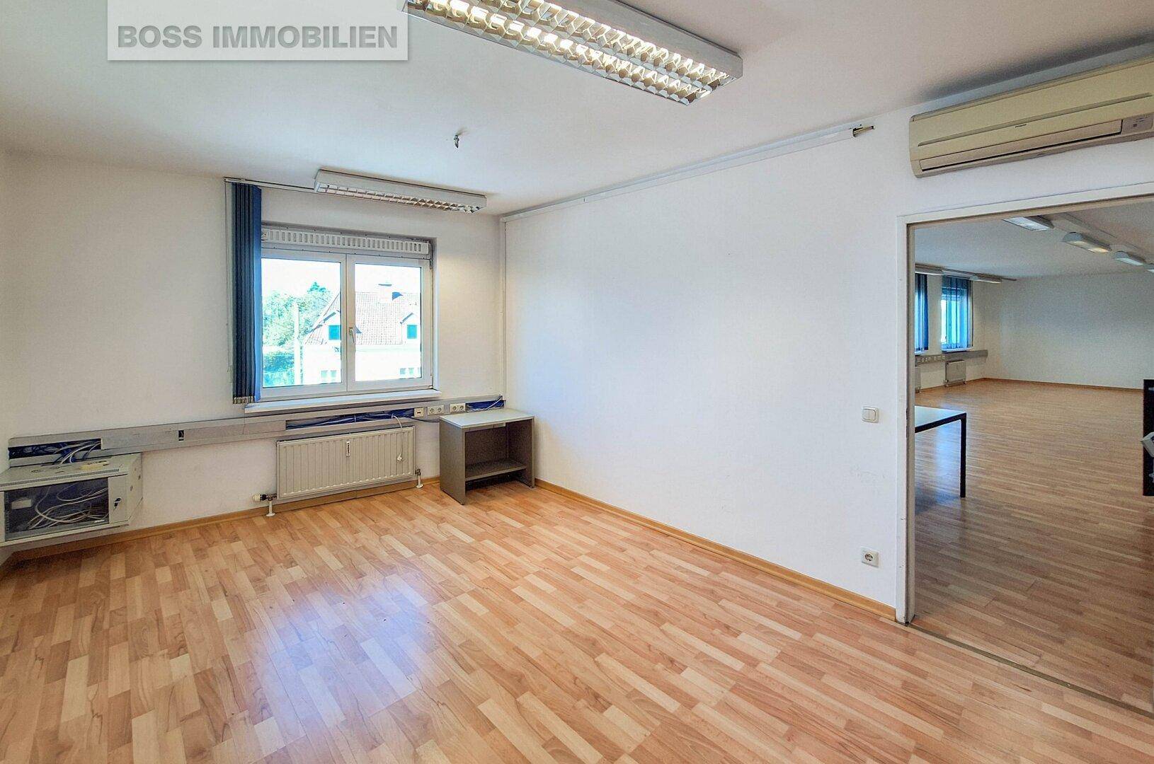 Büro mit Klimaanlage, Laminatboden und einem Durchgang zum größeren Hauptbüro.