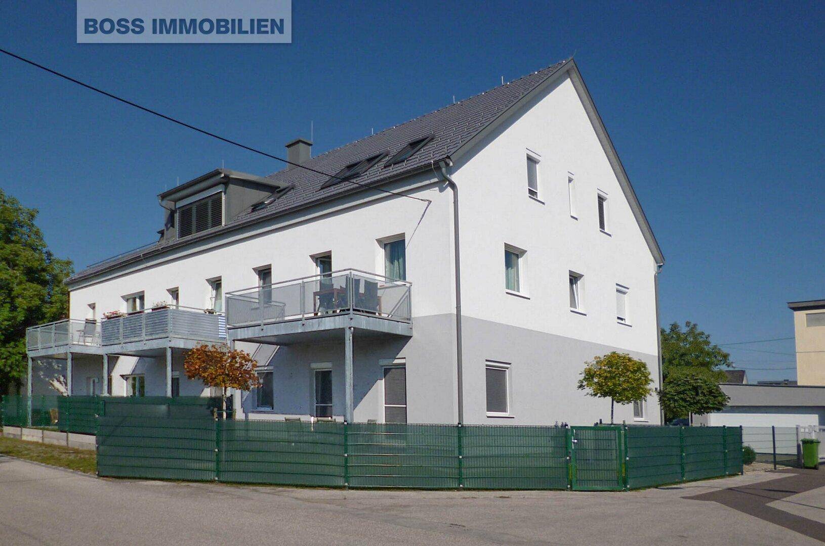 Ansicht eines Mehrfamilienhauses mit weißer Fassade, Balkonen und grüner Umzäunung an einem sonnigen Tag.