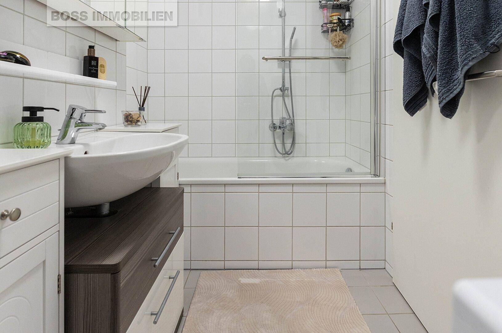 Helles Badezimmer mit weißen Fliesen, Waschbecken und Badewanne mit Duschfunktion.