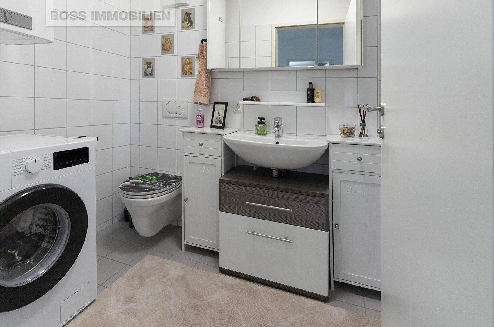 Badezimmer mit weißen Fliesen, Waschbecken, Toilette und integrierter Waschmaschine.