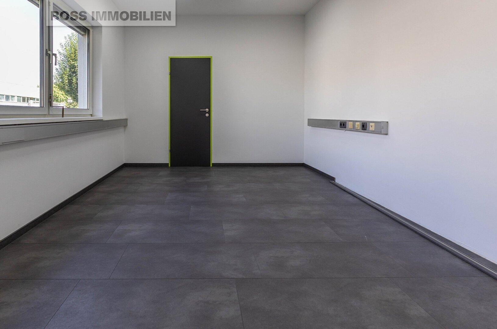 Heller Büroraum mit großem Fenster, dunklem Fliesenboden und einer schwarzen Tür.