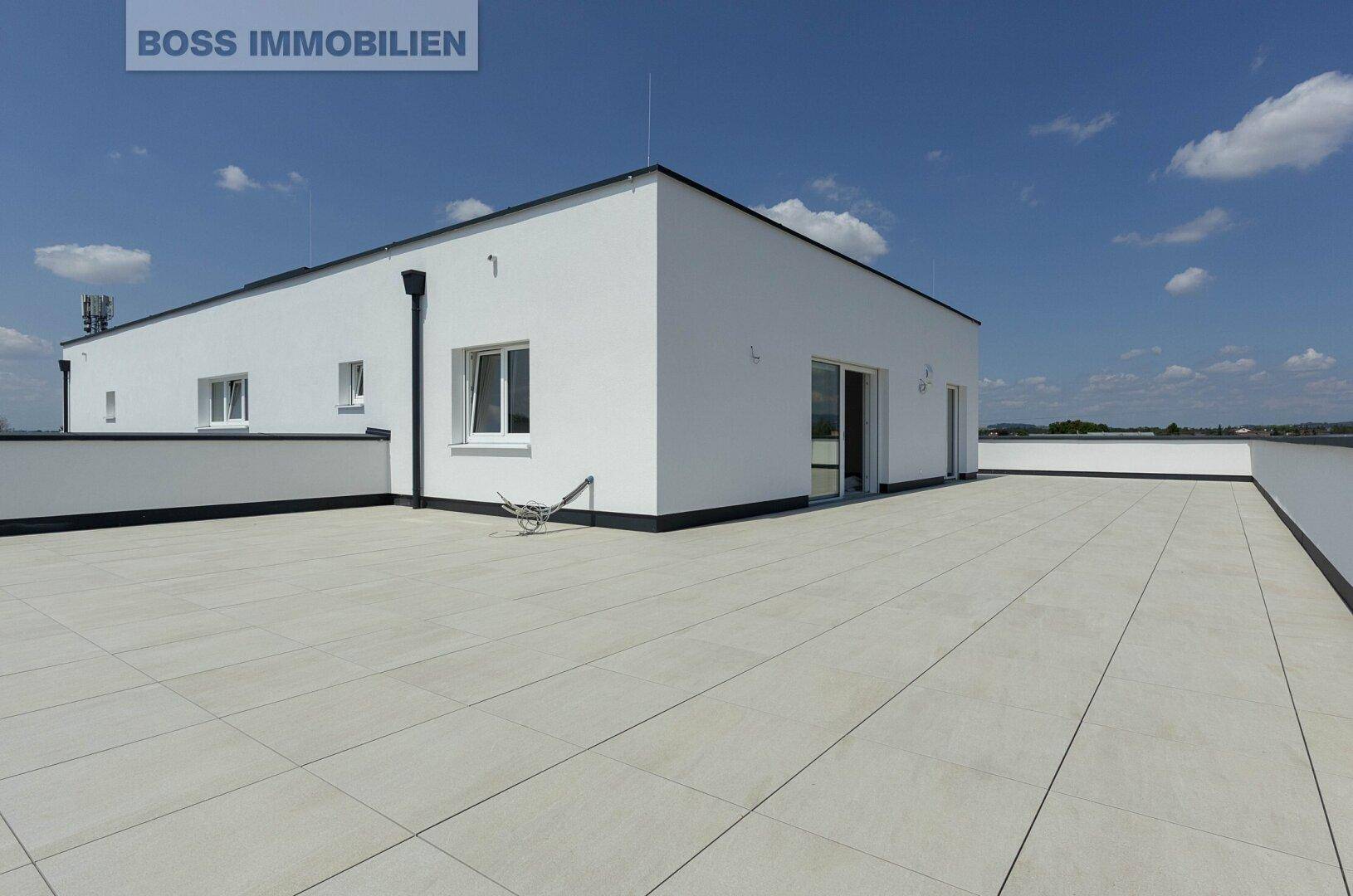 Moderne Architektur mit großer, heller Dachterrasse und weißer Fassade unter blauem Himmel.