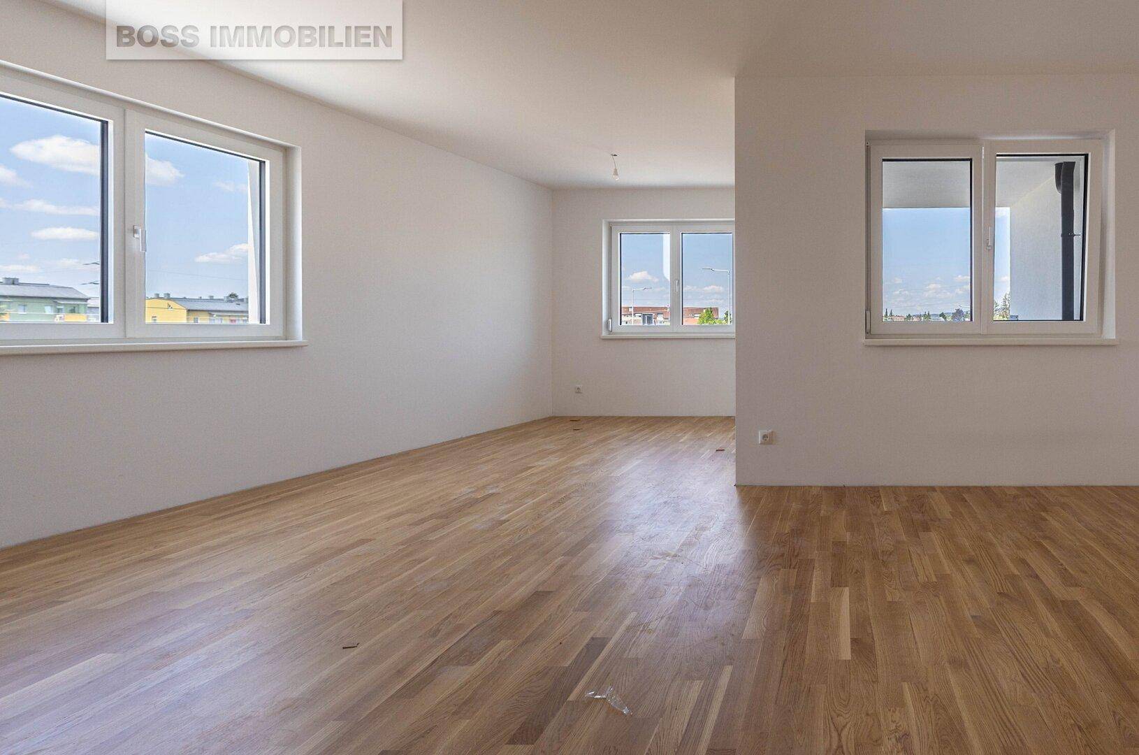 Heller, leerer Raum mit Holzboden und mehreren Fenstern, der viel Tageslicht hereinlässt.