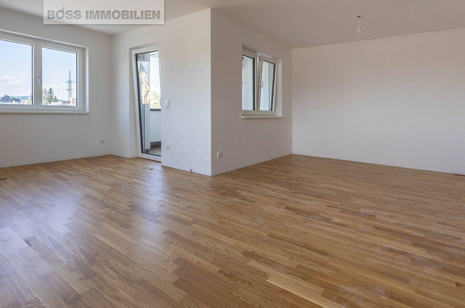 Geräumiges, leeres Wohnzimmer mit Holzboden, großen Fenstern und Zugang zu einem Balkon.