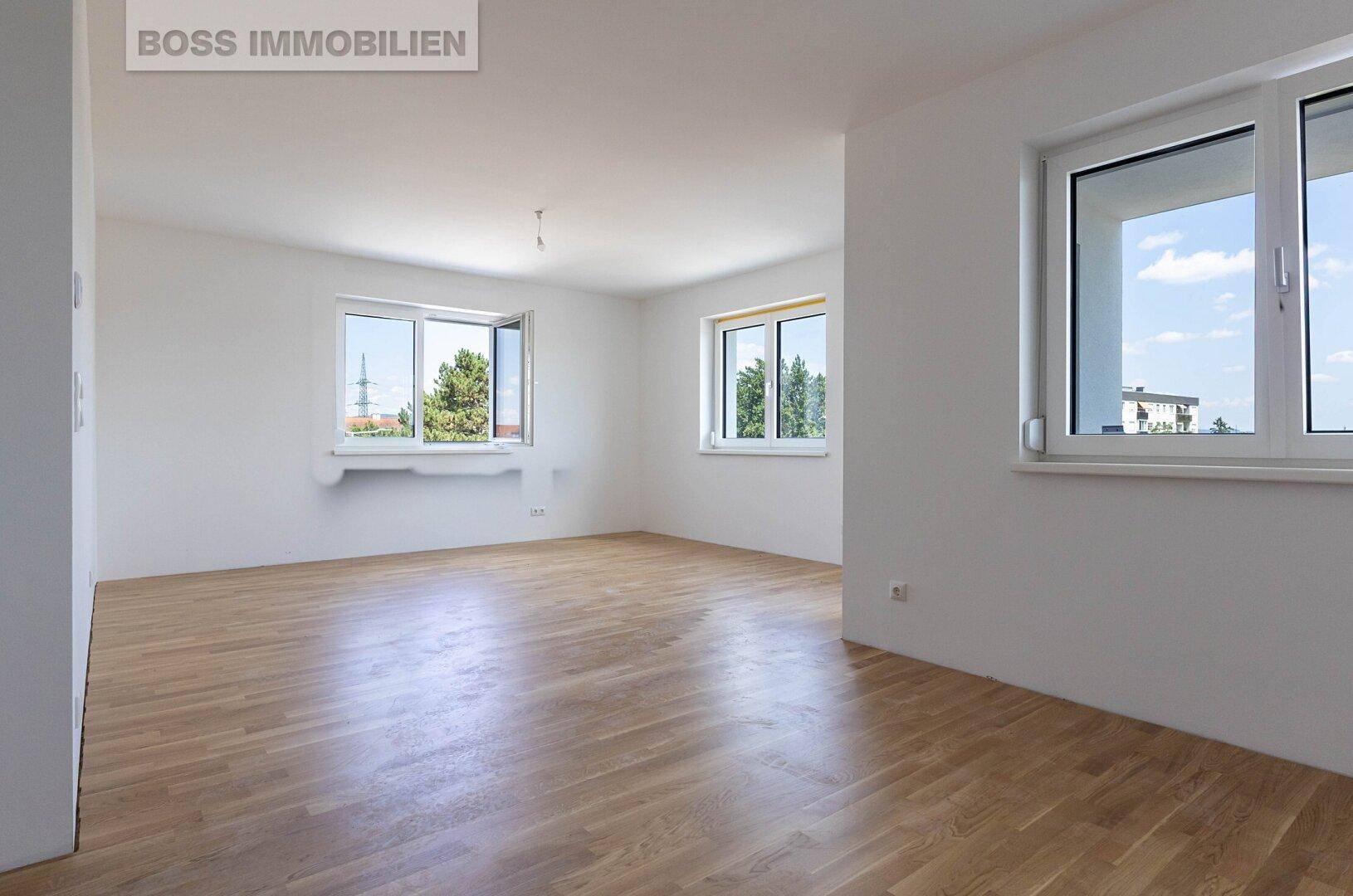 Heller, leerer Wohnraum mit Holzboden und mehreren Fenstern, die viel Tageslicht hereinlassen.