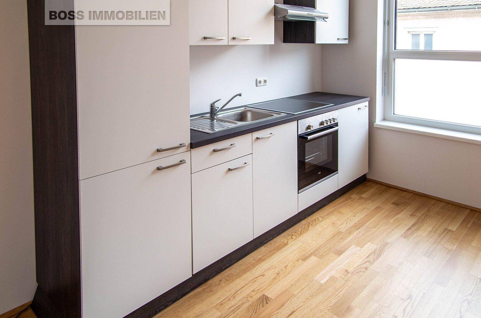 Blick auf die moderne Küche mit Fenster, weißen Schränken, dunkler Arbeitsplatte und hellem Holzfußboden.