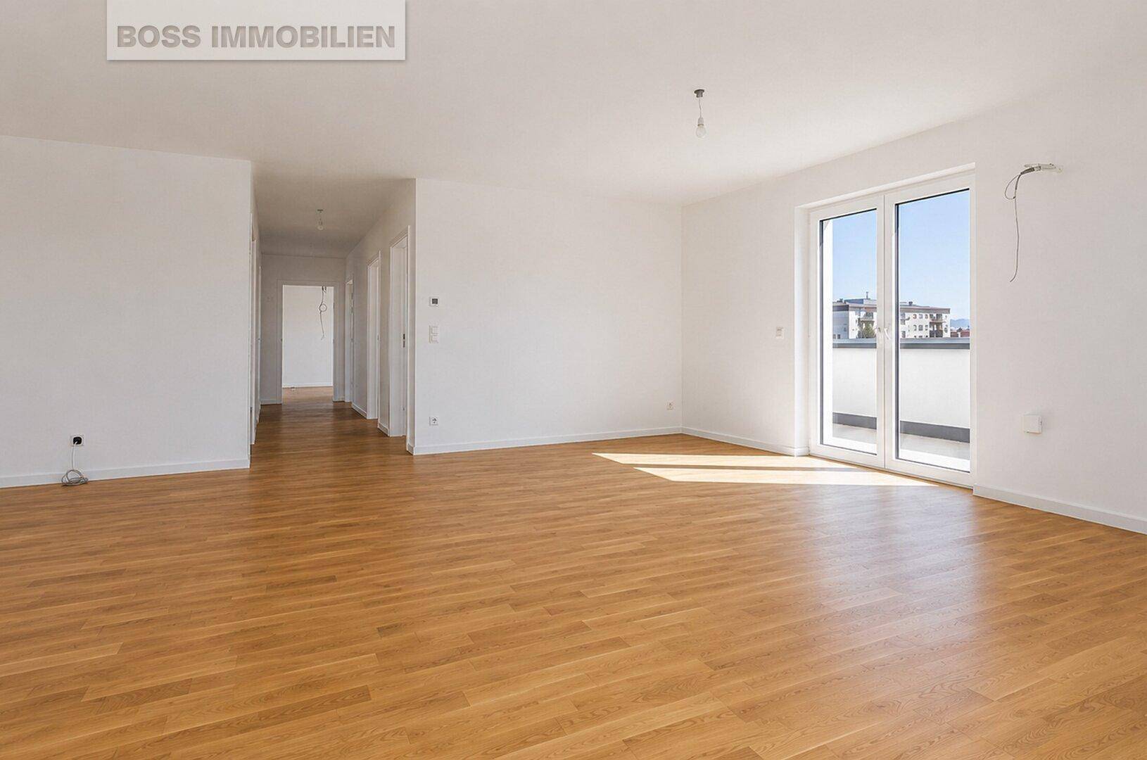Helles, leeres Wohnzimmer mit Parkettboden und Zugang zu einem Balkon.