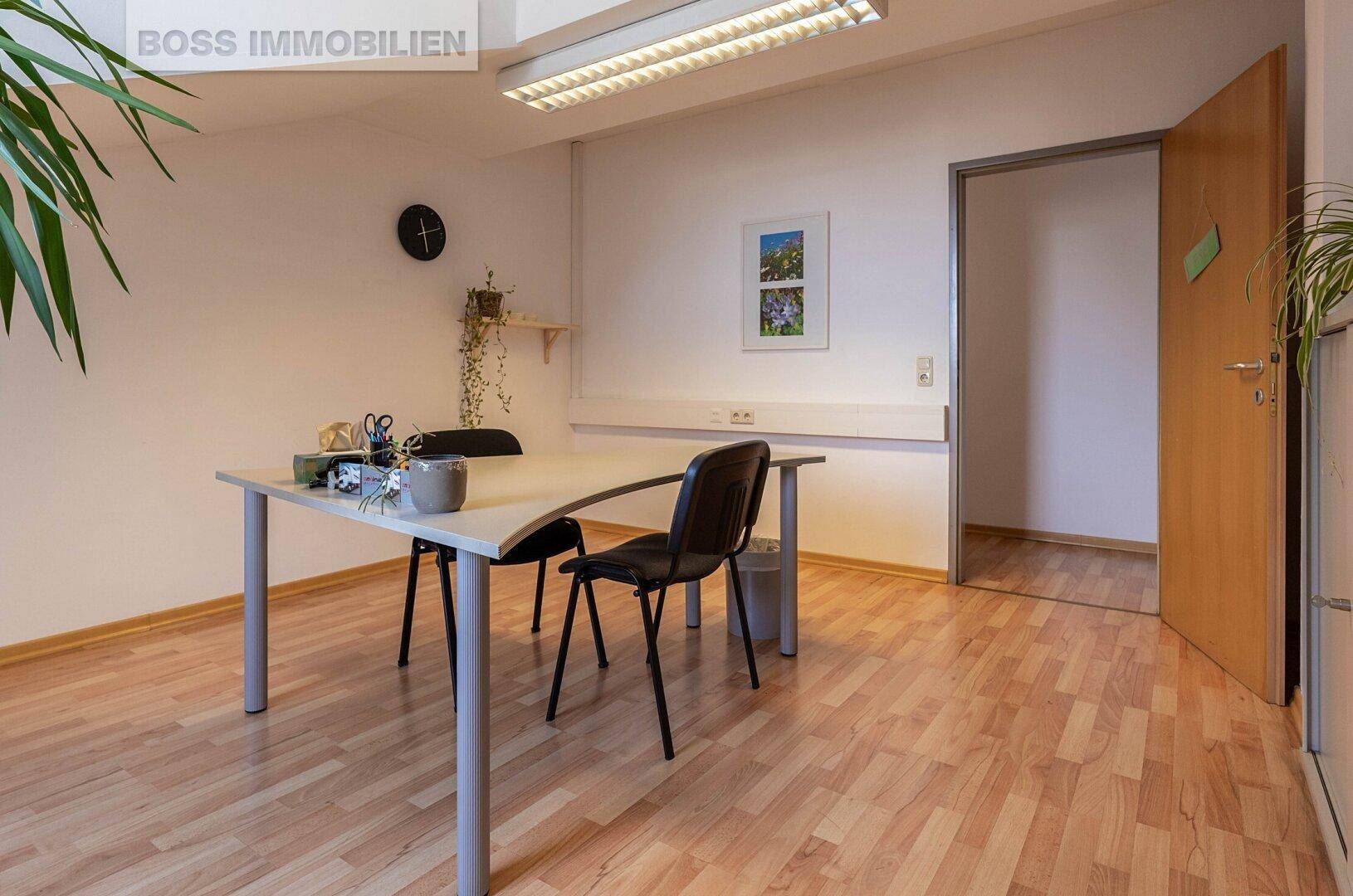 Einzelbüro mit Schreibtisch und Stühlen, hellem Holzfußboden und einer Tür, die zu einem weiteren Raum führt.