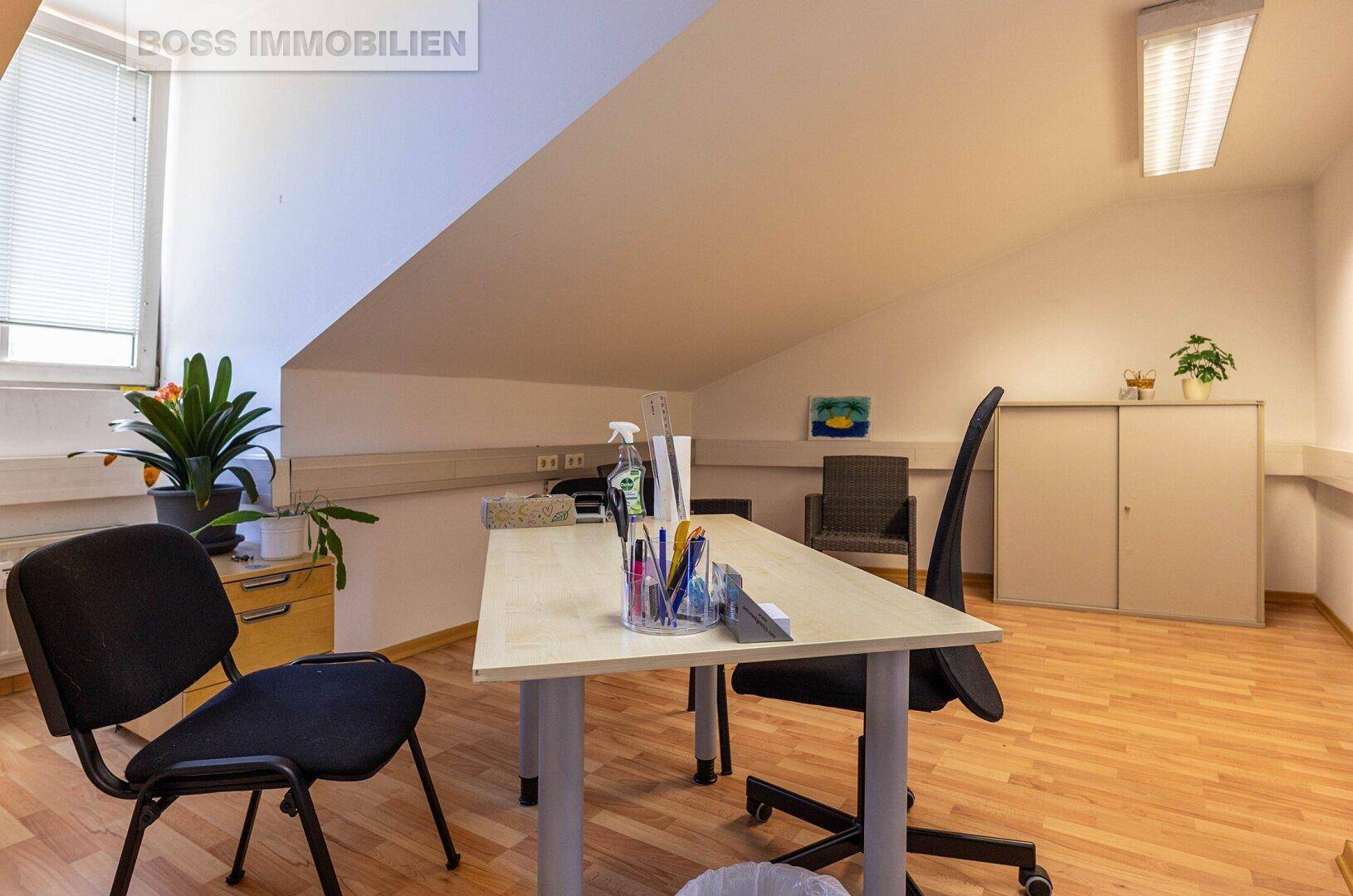 Helles Büro mit Schreibtisch, Bürostühlen, einem Fenster mit Jalousien und einer Dachschräge.
