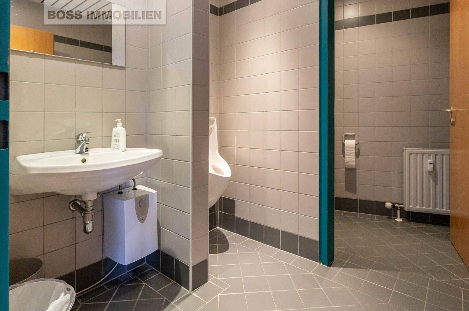 Modern gefliestes Badezimmer mit Waschbecken, Urinal und Heizkörper, in hellgrauen und dunkelgrauen Tönen.