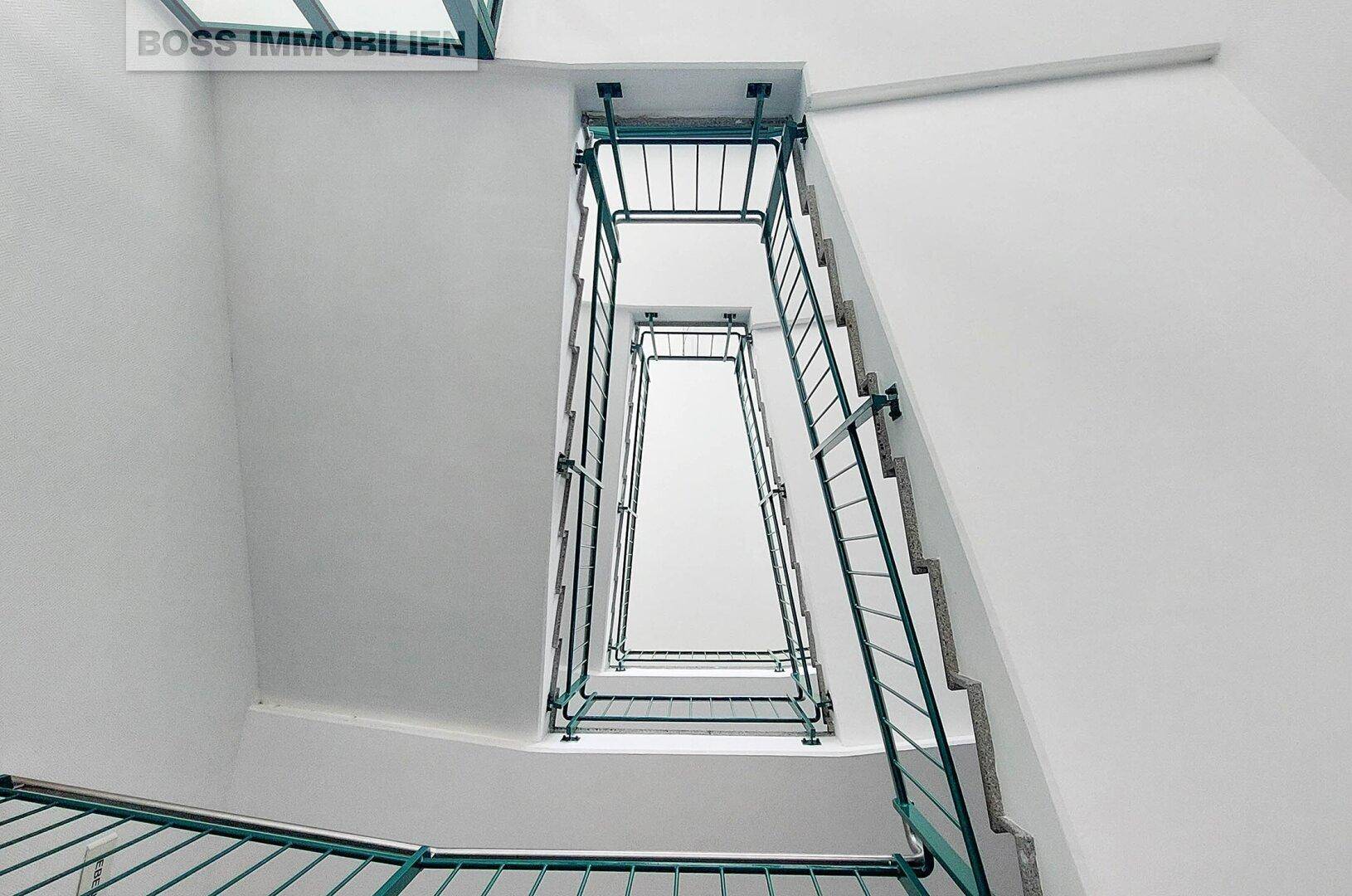Treppenhaus mit grünen Metallgeländern und hellen Wänden, das sich über mehrere Stockwerke erstreckt.