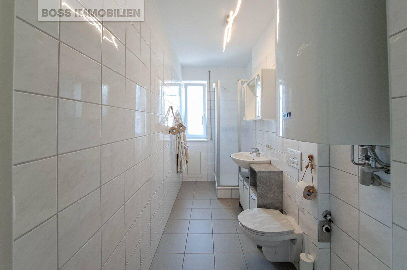 Modernes Badezimmer mit weißen Fliesen, Dusche, WC, Waschbecken und Fenster für Tageslicht.