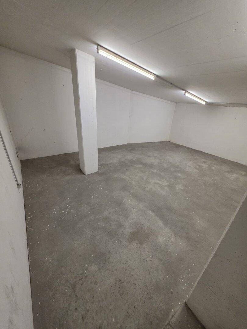 Leerer Kellerraum mit Betonboden, weißen Wänden und einer tragenden Säule, beleuchtet von Deckenleuchten.