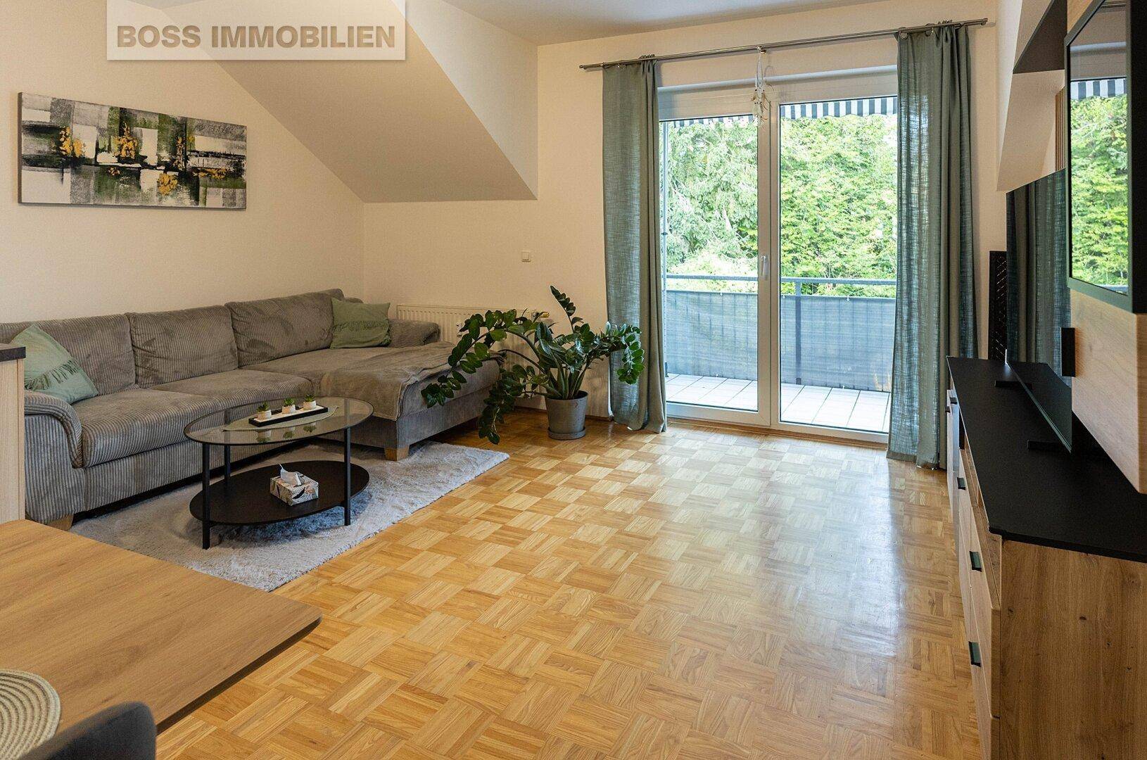 Helles Wohnzimmer mit Parkettboden, Sofa und Zugang zu einem Balkon mit Blick ins Grüne.