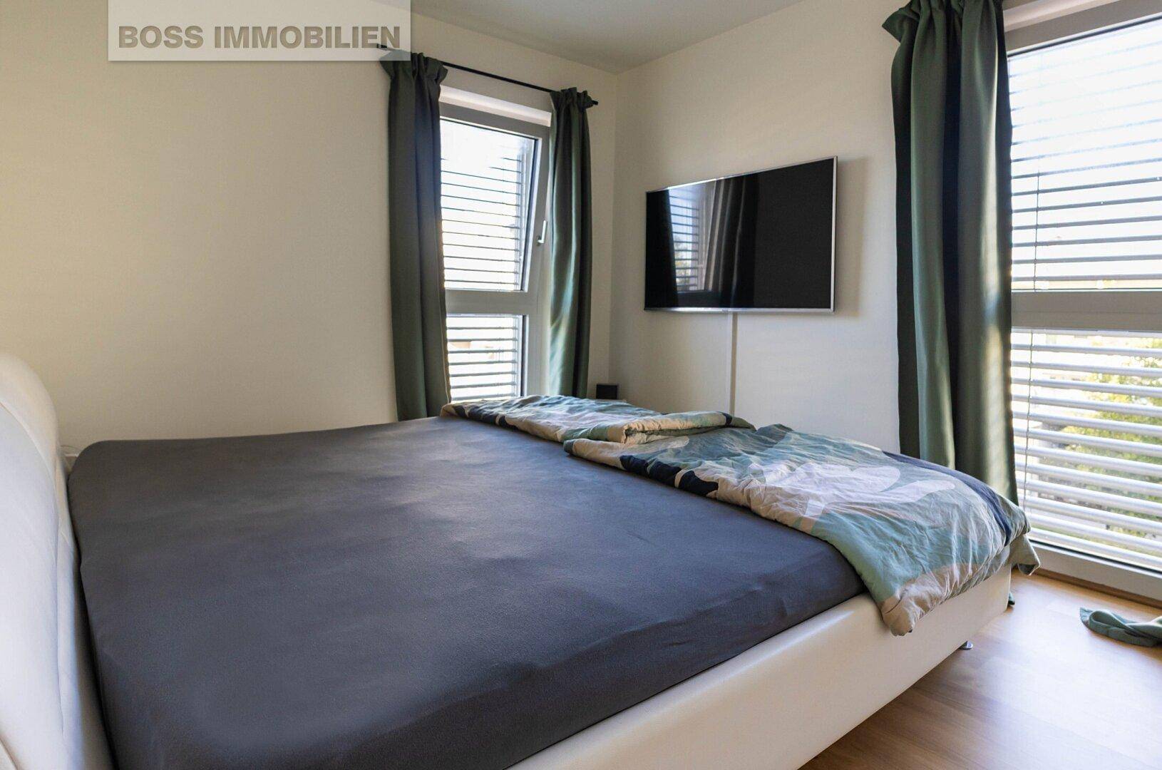 Schlafzimmer mit Doppelbett, Wandfernseher und zwei großen Fenstern mit Jalousien.
