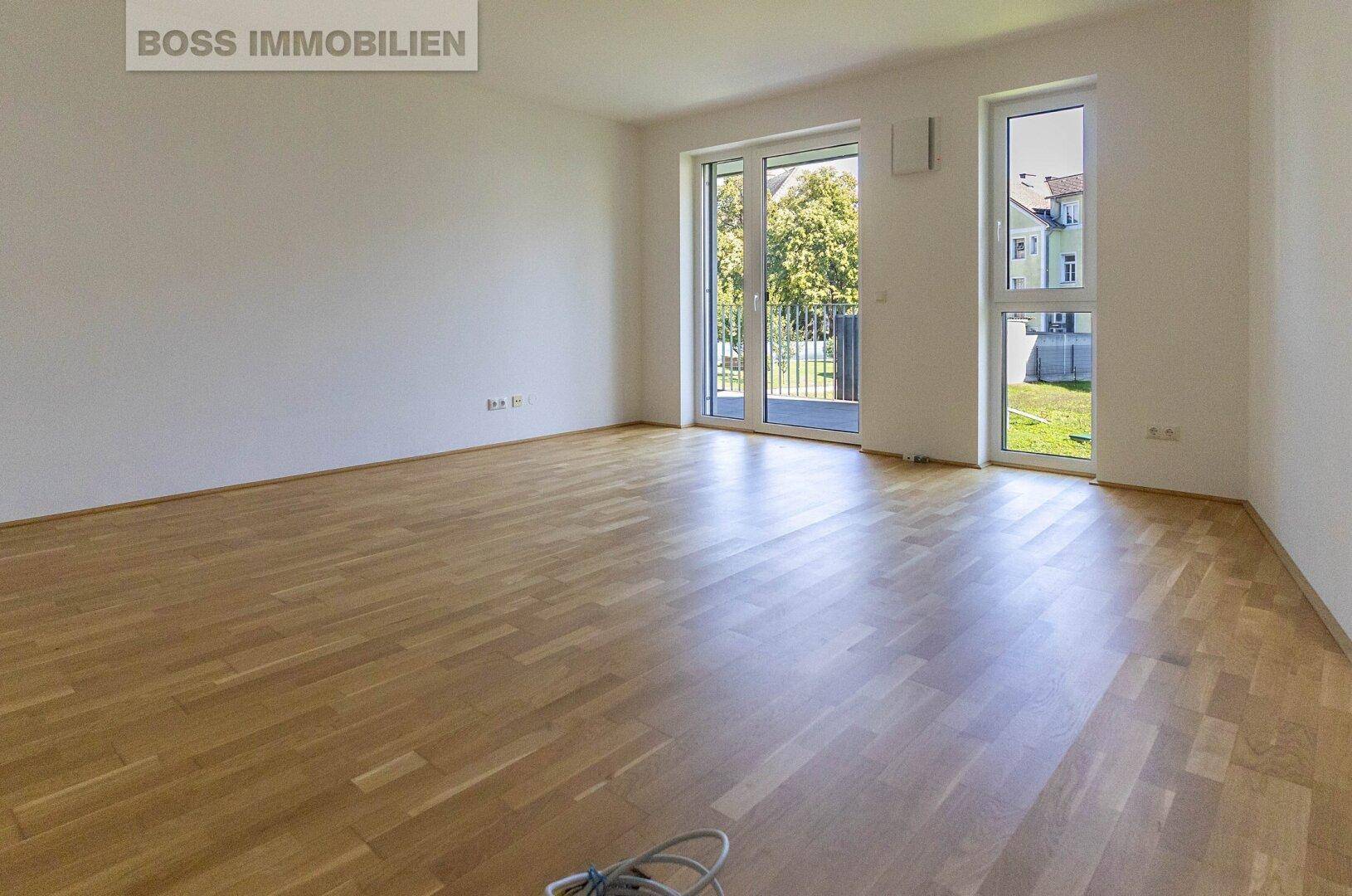 Helles, leeres Wohnzimmer mit Parkettboden und Zugang zu einem Balkon.
