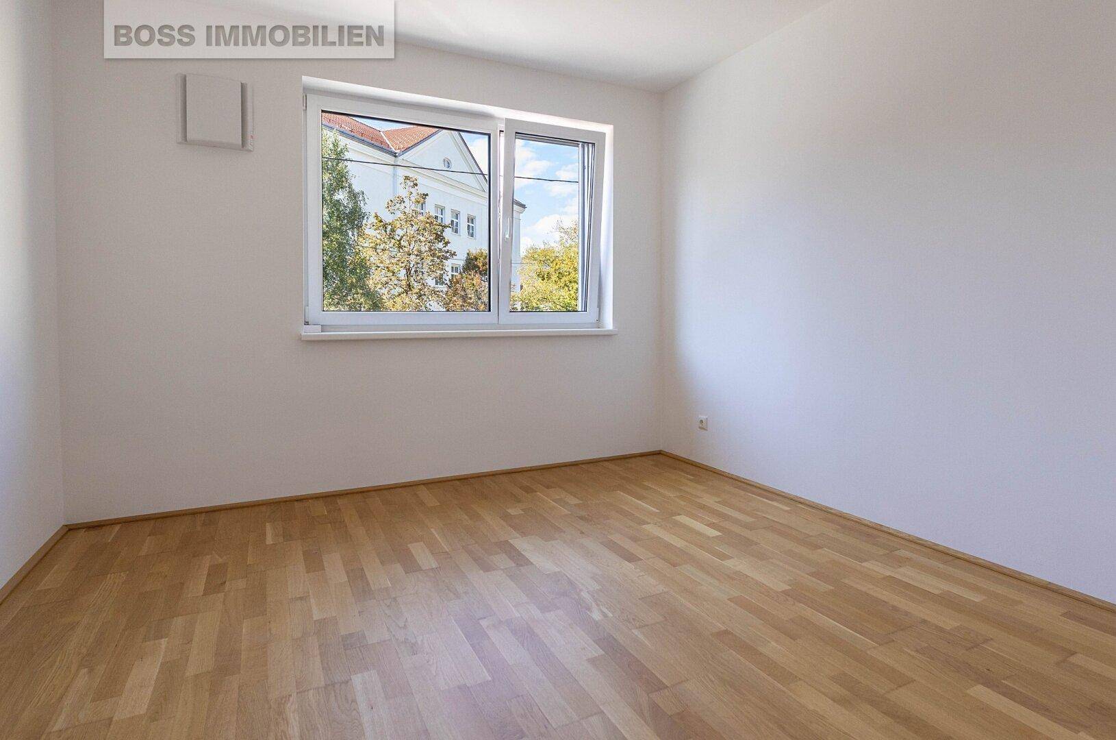 Helles, leeres Schlafzimmer mit Parkettboden und Fenster mit Blick ins Grüne.