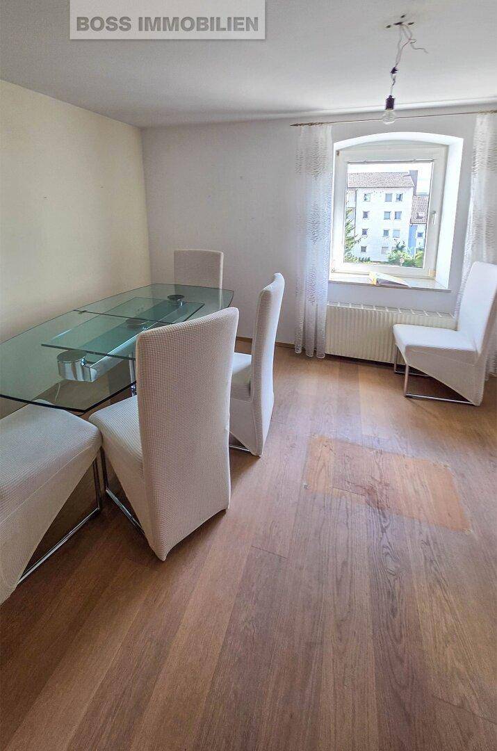 Essbereich mit Holzboden, modernem Glastisch und weißen Stühlen neben einem Fenster.
