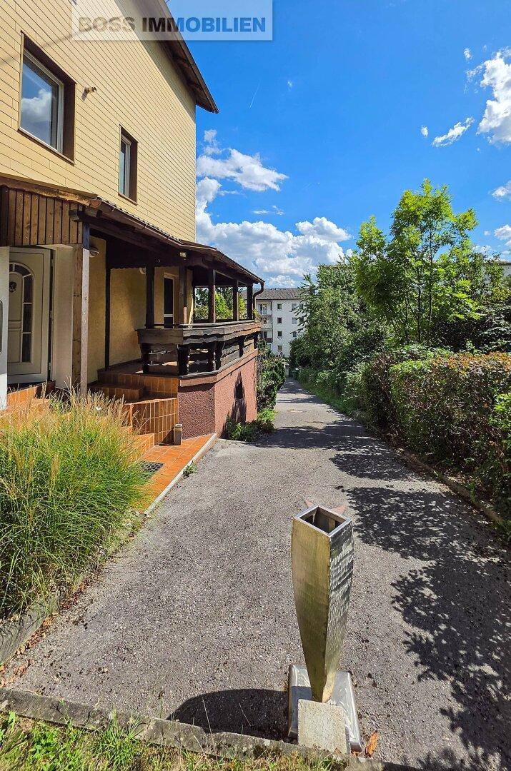 Zufahrt zum Haus mit grüner Bepflanzung und einem modernen Standelement.