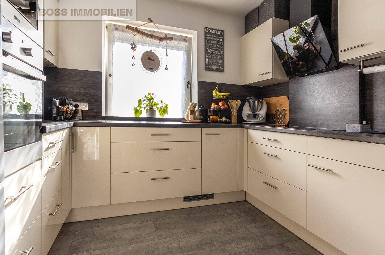 Moderne Küche mit hellen Hochglanzfronten, dunkler Arbeitsplatte und Fenster.