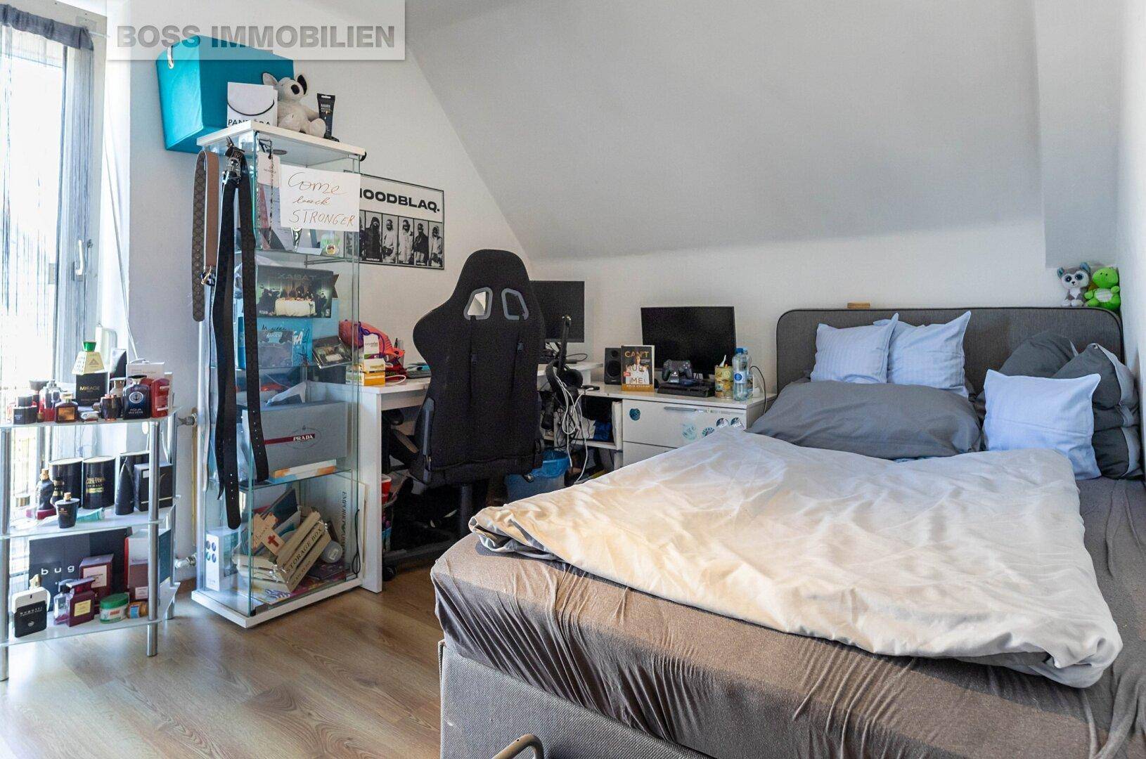Schlafzimmer mit Bett, Schreibtisch und Glasvitrine unter Dachschräge.