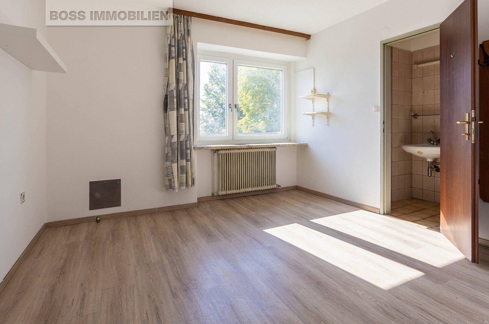 Helles Schlafzimmer mit Laminatboden, großem Fenster und Zugang zu einem kleinen Badezimmer.