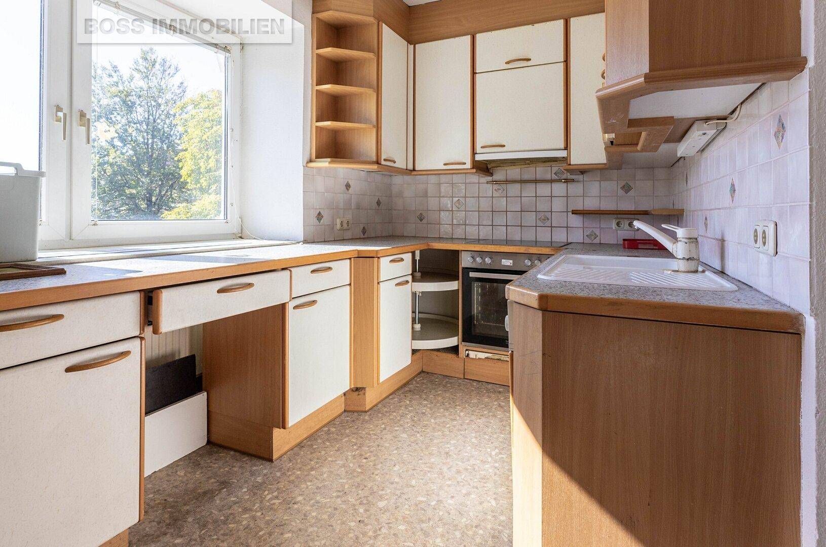 Ältere Küche mit Holzschränken, weißen Fronten, Spüle und Backofen, mit Fensterblick.