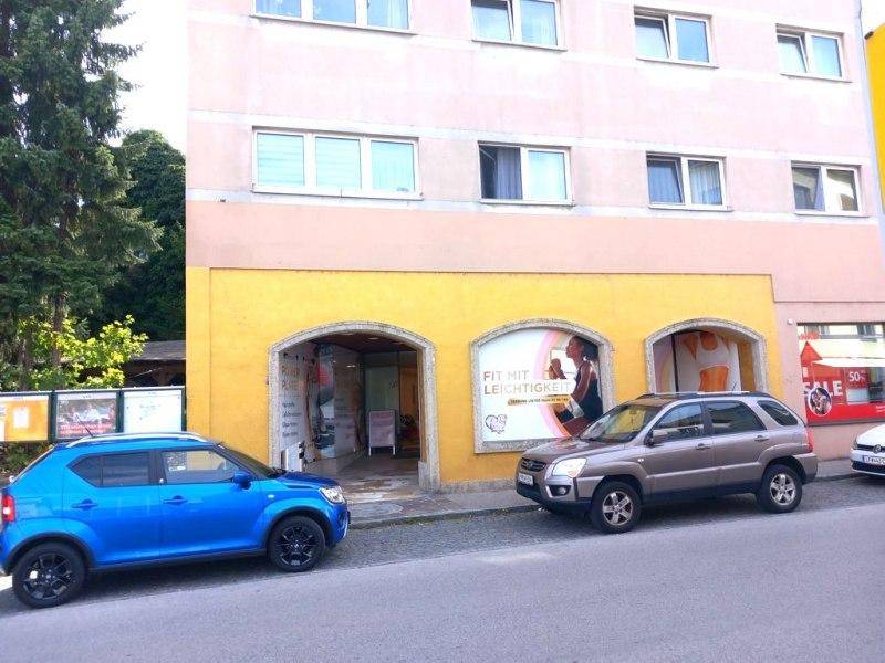 Gebäudeansicht mit Geschäftsräumen im Erdgeschoss und parkenden Autos auf der Straße.