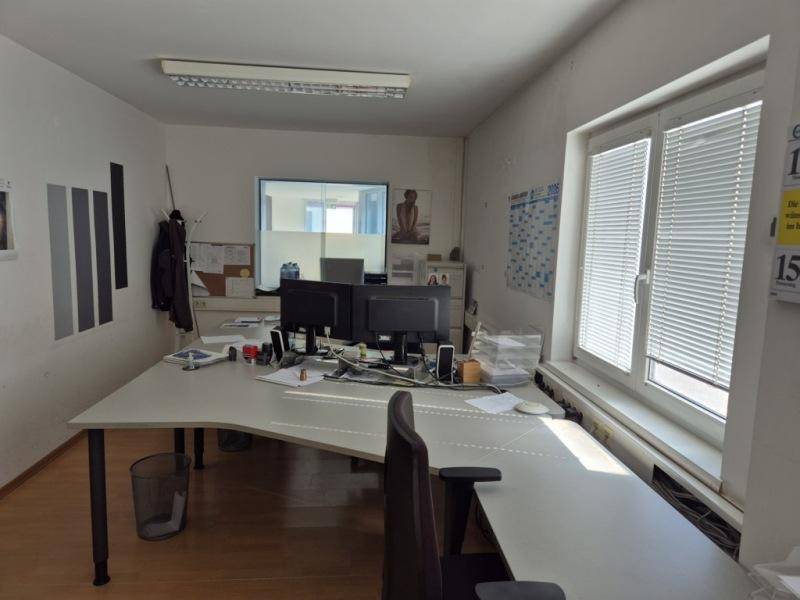 Einzelbüro mit zwei Schreibtischen, Computern und einem Fenster mit Jalousien.