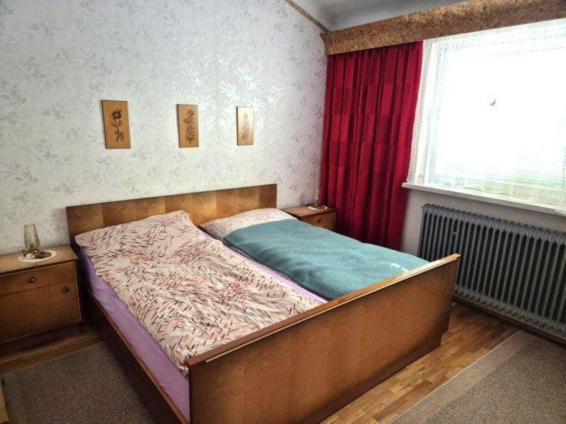 Schlafzimmer mit Doppelbett, Nachttischen und rotem Vorhang, mit gemusterter Tapete.